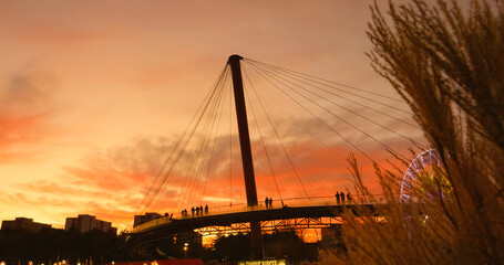 Obraz premium Pedestrians casting dark silhouettes while crossing urban bridge during golden sunset, revealing tranquil cityscape with dramatic sky and architectural elements