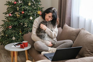 Smiling woman sits comfortably on sofa next to Christmas tree cuddling fluffy grey cat while laptop is open nearby. Cozy winter moments, animal companionship, relaxation at home