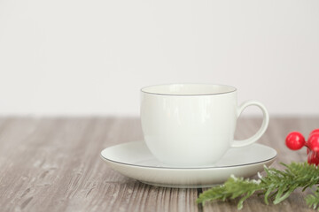 A white porcelain cup with a saucer on a wooden background, decorated with a spruce branch and red berries. Perfect for Christmas and New Year's mood.