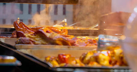 Street food vendor using tongs to serve hot sausages, roasted meat and potatoes on a food stall