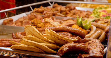 Grilled sausages, flatbreads, and various grilled meats are displayed on a metal tray, ready to be served at a bustling street food market