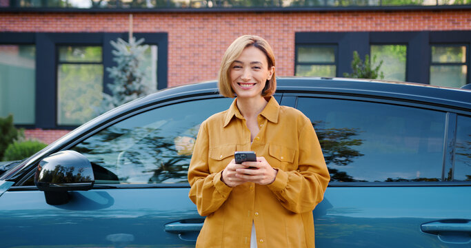 Smiling businesswoman in casual clothes uses a smartphone while standing next to her electric car, looking at the camera and smiling. Modern lifestyle and eco-friendly transport concept. - Powered by Adobe