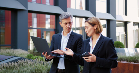 Handsome man in suit and business woman with laptop and smartphone in hands walking and talking at urban area. Business colleagues discussing working issues outdoors.