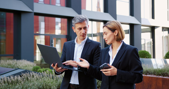 Handsome man in suit and business woman with laptop and smartphone in hands walking and talking at urban area. Business colleagues discussing working issues outdoors. - Powered by Adobe