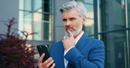 Senior grey-haired bearded businessman in a formal suit standing outdoors, using smartphone to browse social media and manage work tasks.
