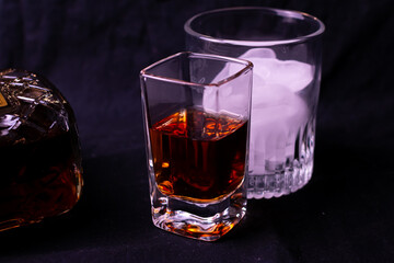 Liquor in a square shot glass beside a cup of ice in a cocktail glass. The bottle of brown liquor is visible partially in the photograph which is set on a black background. 