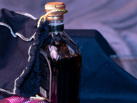 A bottle of brown liquor with a gold top. There is a cloth bag attached to the top. In the lower corner of the photography there are pink small shot glasses. The background is black and grey. 