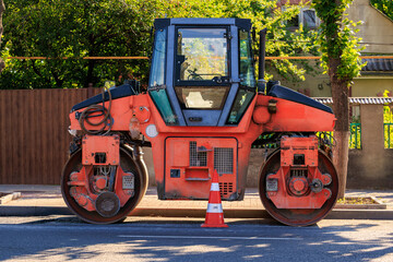 Orange road roller on urban street with safety cone and greenery in background
