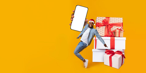 Person dressed in casual attire jumps joyfully beside a stack of beautifully wrapped gifts. Bright yellow background adds to the cheerful holiday atmosphere, while a mobile device is held prominently.