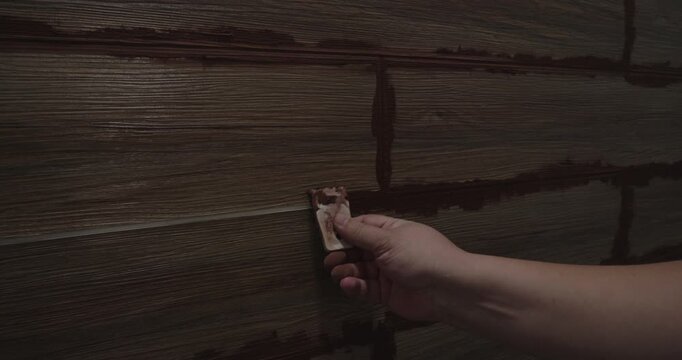 Tiler applying brown grout to the joints between wood-look ceramic tiles on a wall, using a small rubber spatula to fill the gaps during a home renovation project and finishing process.