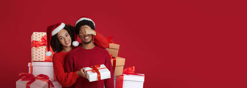 A joyful couple stands together against a bright red background, surrounded by wrapped gifts. They wear Santa hats and festive attire while sharing a playful moment, enhancing the holiday spirit.