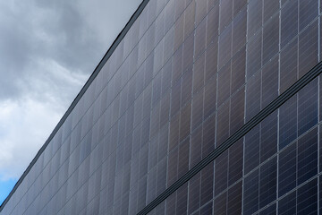 Solar panels cover a large building wall under a cloudy sky in a modern urban setting