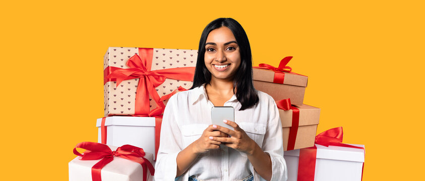 A cheerful individual stands surrounded by beautifully wrapped gifts, holding a smartphone. The background features a bright yellow color, adding energy to the festive scene. - Powered by Adobe