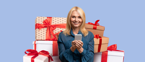 A cheerful woman is smiling while holding a smartphone. She is surrounded by various gift boxes...