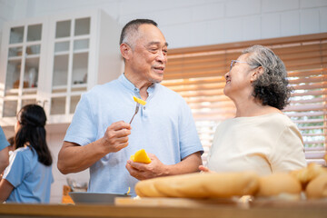 Asian elderly couple enjoying sweet kitchen moments while preparing food. The husband lovingly feeds his wife fruit, reflecting care, companionship, and joyful aging in a warm home.