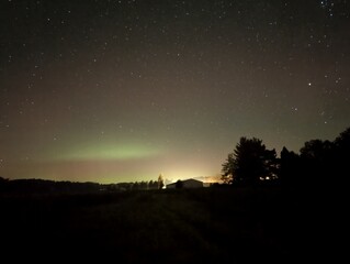 Night Sky with faint aurora and illuminated horizon. A dark field stretches towards the glowing horizon, with trees silhouetted against the dark sky