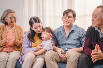 Asian multigenerational family sitting together on the sofa, interacting with toddler in a loving,...