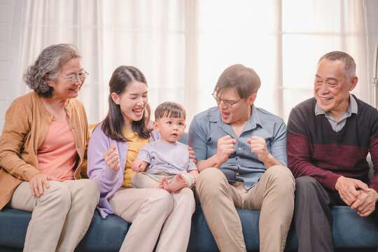 Asian multigenerational family sitting together on the sofa, interacting with toddler in a loving, joyful moment at home, representing close family bonds, parenting, and emotional connection.