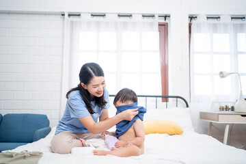 Asian mother dressing her cheerful toddler after bath, while the baby holds a lotion bottle. A joyful moment of parenting, baby care, and cozy family bonding in daily routine.