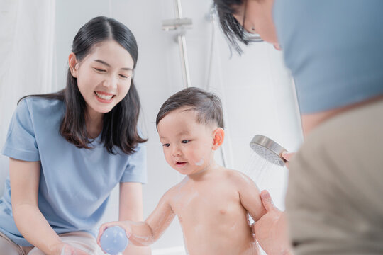 Asian parents giving their toddler a joyful bath, using a handheld shower in a modern bathroom. A happy family moment showcasing love, hygiene, and playful parenting routine.
