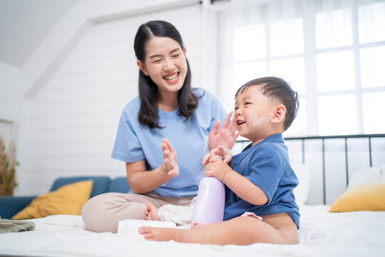 EN: Asian mother smiling while dressing her happy toddler after bath. The baby holds a lotion bottle, capturing a warm family bonding and baby skincare routine in a cozy bedroom.