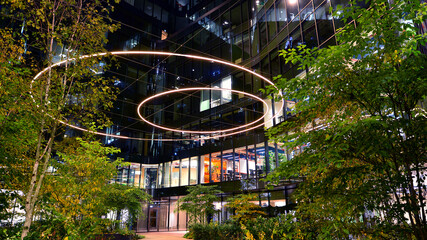 Modern office building in city center illuminated at night. Green trees near a modern office building with a glass facade. Rows of lit windows against the architectural grid of a modern facade.