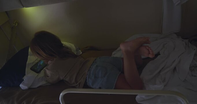 A child enjoys watching a movie on a tablet during a train ride. A little girl lies on her bunk bed in a dark sleeper car compartment, kicking her legs during the train ride.