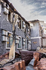 Ground level view of fire damaged building blackened brick walls broken windows twisted metal debris and intact AC units contrast with collapsed roof and scorched upper facade.