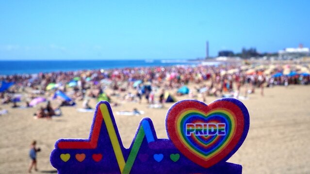 Rainbow heart symbol with PRIDE text, representing summer celebration and LGBTQ+ holiday, standing on a sunny beach full of people enjoying the warmth and freedom - Powered by Adobe
