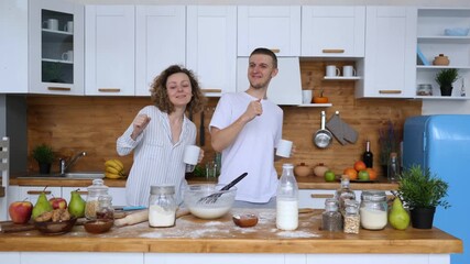 Joyful young couple in pajamas dancing together in the kitchen while preparing a delicious and healthy breakfast, enjoying their morning routine and having fun with food ingredients on the table - Powered by Adobe