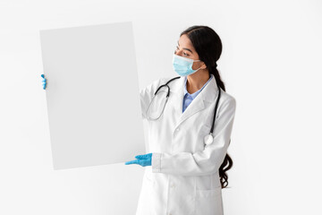 Millennial Indian woman doctor poses with a large blank banner while wearing a protective mask and coat. She stands against a plain white background, ready for health-related announcements.
