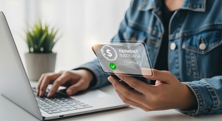 A young Asian woman in a denim jacket smiles at her smartphone, confirming a successful online payment while working on a laptop.