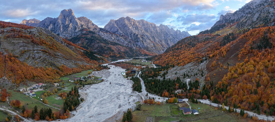 Panoramic sunrise view of Valbona Valley with snow-dusted peaks, autumn forests, and the wide riverbed below.