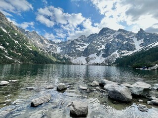 A stunning lake reflects the towering snow-capped mountains and cloudy sky. Rocky shores create a serene atmosphere in this natural setting, perfect for exploration and relaxation.