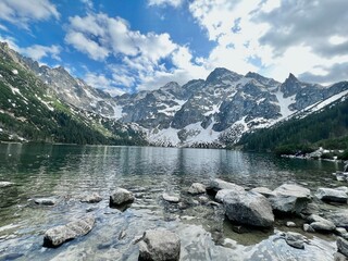 Lush green forests surround a tranquil lake reflecting the vivid blue sky and dramatic snow-capped mountains. The rocky shore adds texture to this stunning natural scene.