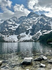 Snow-covered mountains rise majestically above a calm lake, with rocky shores and clouds floating softly in the sky on a peaceful day in nature.