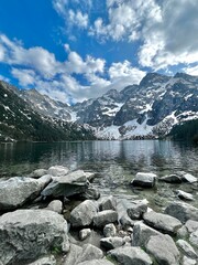A serene mountain lake reflects the towering snow-covered peaks on a clear day. Lush greenery surrounds the water, creating a peaceful atmosphere.