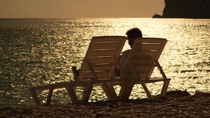 Sunset beach relaxation, Young man sits on a beach chair watching the beautiful golden sunset over the calm sea - Powered by Adobe