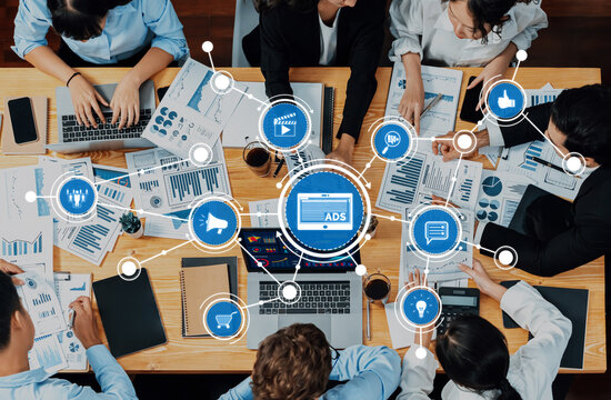 A dynamic business meeting showcasing a diverse team analyzing marketing data and strategies using ADS technology. The wooden table is filled with charts and reports. Amity - Powered by Adobe