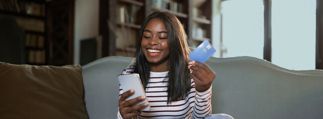 A young woman sits comfortably on a couch, smiling as she looks at her smartphone while holding a credit card. Sunlight pours through large windows, creating a warm atmosphere.