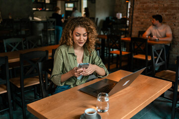 Young woman using smartphone and laptop in a cozy cafe setting during the day