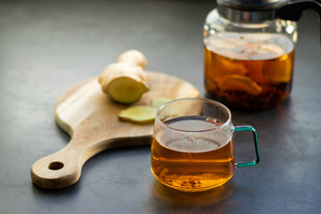 A glas cup of hot tea with fresh ginger and glass tea pot standing on greys kitchen table 