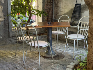 Wooden table and metal chairs under tree shade in outdoor cafe. Peaceful corner, harmony of nature and urban comfort in timeless style.