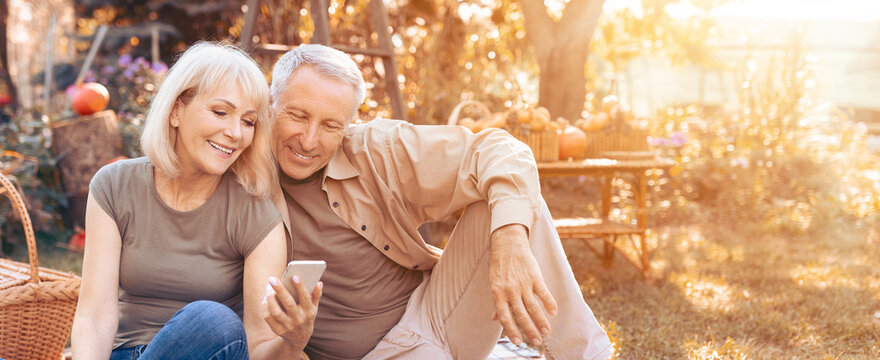 A smiling couple sits on a blanket in the garden, looking at a smartphone. The warm sunlight creates a cozy atmosphere, surrounded by colorful autumn decorations.