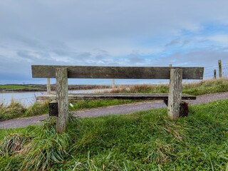 Old weathered wooden bench by a grassy coastal path overlooking the sea and cloudy blue sky.