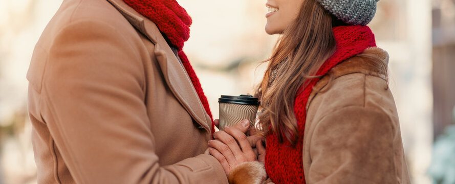 A happy couple stands close together in a winter setting, smiling at each other while holding coffee cups. They are bundled in warm coats and scarves, surrounded by a festive atmosphere.