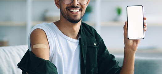 A young man smiles while holding a smartphone in his hand, displaying his vaccination record. He has a bandage on his arm and is seated in a bright, stylish room.