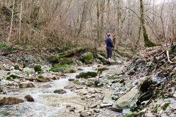 A man stands in a dry riverbed surrounded by bare trees, gazing into the distance with contemplative solitude.