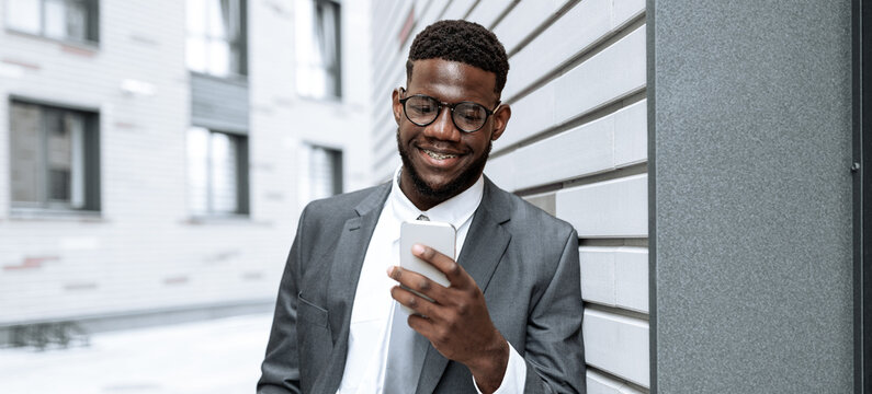 A man dressed in a suit with glasses smiles while looking at his smartphone. He stands leaning against a sleek building with neutral tones in an urban setting during daylight hours.