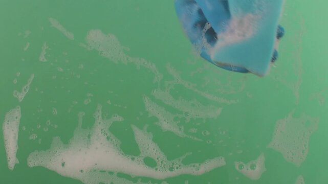 A hand in a rubber glove with a sponge washes the surface. Hands in rubber gloves hold the sponge and scrub the surface. The sponge is covered in sudsy cleanser on a chroma key.